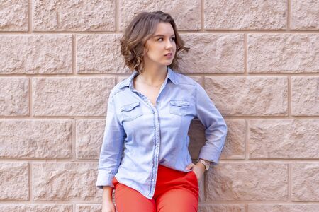 female portrait. beautiful young girl stands on a background of a beige brick wall with stucco. red pants and a denim shirt. beige backgroundの写真素材