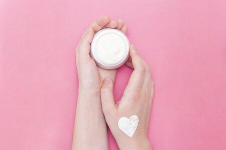 Hands of a beautiful well-groomed woman with a cream jar on a pink textural background. Moisturizer for clean and soft skin in the winter. The heart shape is made of cream. I love the body. Healthcare concept.の写真素材