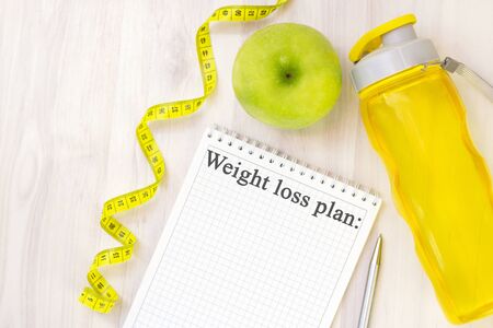 green apple, measuring tape and notebook for writing with the inscription weight loss plan on a light wooden background. preparation for the summer season and the beach, weight loss and sports conceptの写真素材
