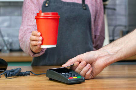 Takeaway coffee. barista holds out a red paper glass with a drink, the buyer pays with a card
