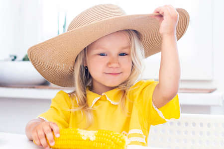Child girl in a straw hat in yellow clothes eats corn, summer photo. on a light backgroundの写真素材