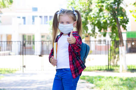 happy schoolgirl in a protective mask with a backpack and a textbook in her hands. in a white T-shirt and a plaid shirtの写真素材
