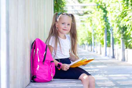 schoolgirl sits on a bench in the street with a backpack and reads a textbook, book. Back to school. A cute schoolgirl with long ponytails.の写真素材
