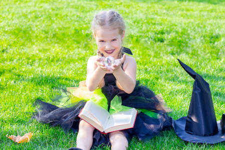 girl in a witch costume for the Halloween holiday. Placard with the inscription: Halloween. Happy girl sitting reads a spell from a bookの写真素材