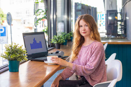 A young woman drinks coffee and works on a laptop in a cafe.