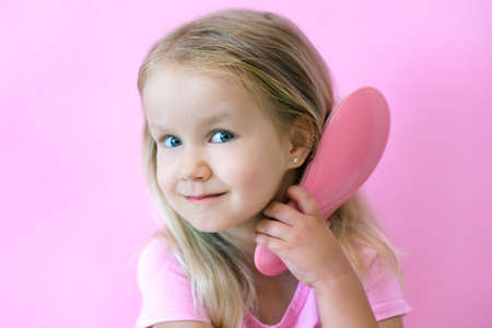 Happy little girl combing her hair. Beauty and childhood concept. Girl on a pink isolated background combing her hair with a pink comb her blonde hairの写真素材