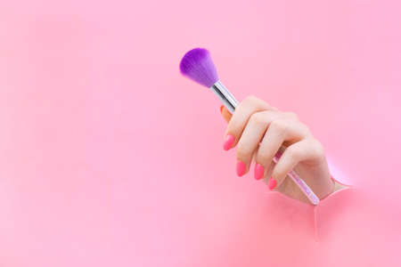 Brush for applying makeup, brush for manicure in the hand of a young woman on a pink background from a hole. copy spaceの写真素材