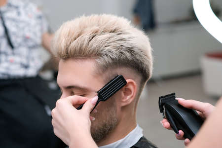haircut process of blond young man with hair trimmer in armchair in barbershop salon, barbershop concept for men and boysの写真素材