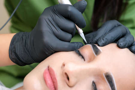 Beautician makes permanent eyebrow makeup on the face of a beautiful young woman