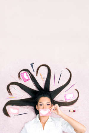 A beautiful young woman lies with long hair, the tools for lamination and care of eyebrows and eyelashes are on a pink background. Young woman holding a patch at her mouth like a smileの写真素材