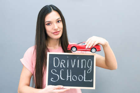 Driving school idea and concept, student driver passed the exam, drivers license, portrait of a beautiful happy young woman, holding a car in her hand. on a gray isolated background. plate with the inscription Driving schoolの写真素材