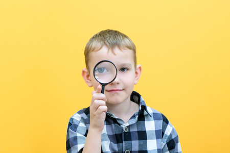 Positive curious schoolboy in casual clothes looks at the camera through a magnifying glass, on a...