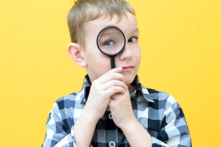 Positive curious schoolboy in casual clothes looks at the camera through a magnifying glass, on a yellow background. surpriseの写真素材