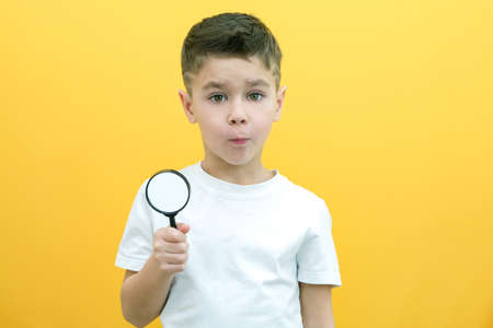 Positive curious schoolboy in casual clothes looks at the camera through a magnifying glass, on a...