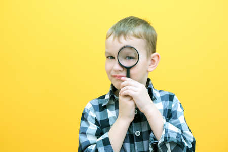 Positive curious schoolboy in casual clothes looks at the camera through a magnifying glass, on a yellow background. surpriseの写真素材