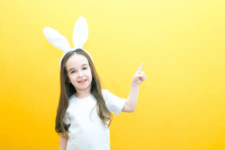cheerful girl with rabbit ears on her head on a yellow background. Funny happy child points fingers at an empty space. copy space for text, mockupの写真素材
