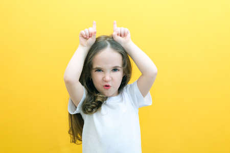 Portrait of cheerful cute little girl showing bull horn gesture, funny kid, joke. shooting indoors, yellow backgroundの写真素材