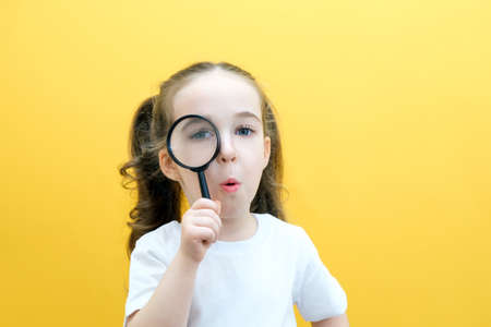 Positive curious schoolgirl in casual clothes looks at the camera through a magnifying glass, on a yellow background. surpriseの写真素材
