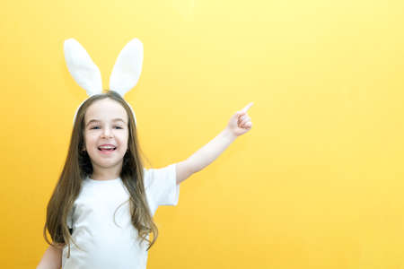 cheerful girl with rabbit ears on her head on a yellow background. Funny happy child points fingers at an empty space. copy space for text, mockupの写真素材