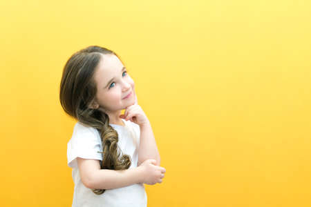 Thoughtful little girl touching observation chin, thoughtful cute daughter making decision, imagining thought, posing isolated over yellow backgroundの写真素材