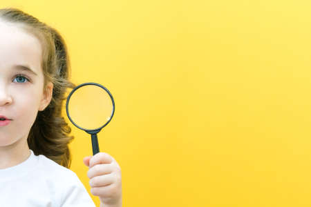 Positive curious schoolgirl in casual clothes looks at the camera through a magnifying glass, on a yellow background. surpriseの写真素材