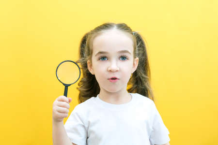 Positive curious schoolgirl in casual clothes looks at the camera through a magnifying glass, on a yellow background. surpriseの写真素材