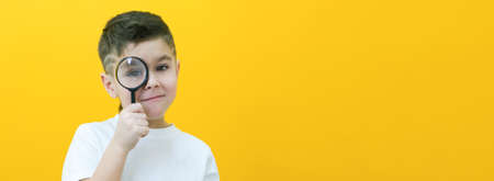 Positive curious schoolboy in casual clothes looks at the camera through a magnifying glass, on a...