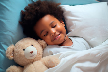 a darkskinned boy sleeps sweetly in bed with a toy bear in his arms under the blanketの素材
