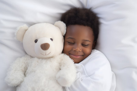 a darkskinned boy sleeps sweetly in bed with a toy bear in his arms under the blanketの素材