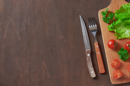 Cherry tomatoes, lettuce, fork and knife, cutting board on a dark wooden background.の写真素材