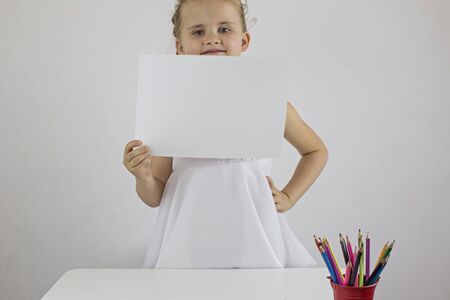 The little girl is standing at the desk and holding a white sheet of paperの写真素材