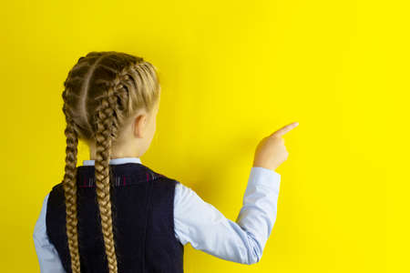 Schoolgirl stands with her back pointing her finger at free empty space for text or advertising, standing isolated on yellow studio backgroundの写真素材