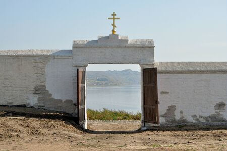 An old white brick fortified wall with traces of repair and with an open gate in the middle, a golden Christian cross above them, through the gate a view of land and water and the other shoreの写真素材