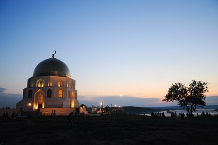 A large mosque with a metal dome beautifully glowing at dusk, standing on a high dark bank of the river against the backdrop of a pink sunset, surrounded by unrecognizable people admiring the viewの写真素材