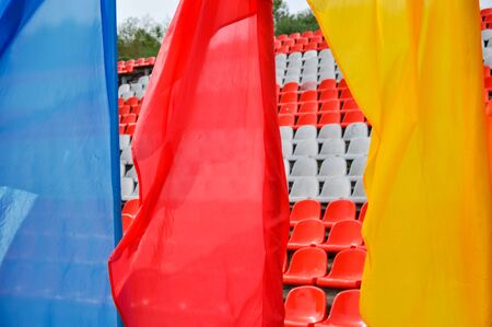 Colorful waving flags and empty seats in the stands of the arena or auditorium. Rows of red and white stadium seats without spectators. Concept of cancellation of mass sports and entertainment eventsの写真素材