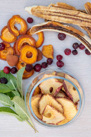 Sliced dried dehydrated apples, apricots and bananas, dried cherries in a glass jar, mint leaves on a light table. The concept of home preparation of dried fruits in a dehydrator.の写真素材