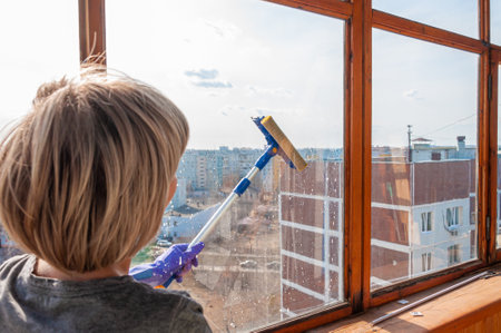 An unrecognizable young woman in blue gloves washes the balcony window from the outside with a special long-handled scraperの写真素材