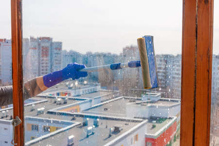 An unrecognizable young woman in blue gloves washes the balcony window from the outside with a...