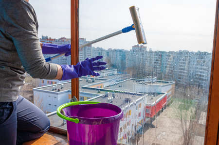 An unrecognizable young woman in blue gloves washes the balcony window from the outside with a special long-handled scraperの写真素材