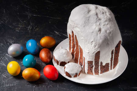 Homemade craft Easter cakes covered with white icing and multicolored painted Easter eggs on plates on a dark background.の写真素材
