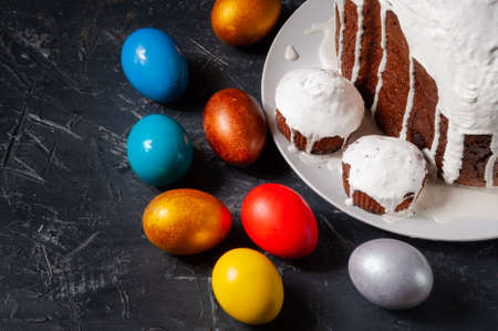 Homemade craft Easter cakes covered with white icing and multicolored painted Easter eggs on plates on a dark background.の写真素材