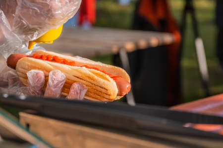 Fresh hot dog with fried sausage, served in an outdoor cafe. Cooking American barbecue food. The chef fills a hot dog with mustard. Barbecue festival in the city park. Street fast food.の写真素材