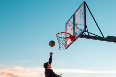 A young basketball player throws a ball into a basketball basket against the blue sky. Transparent plastic basketball shield on the outdoor playground.のeditorial素材