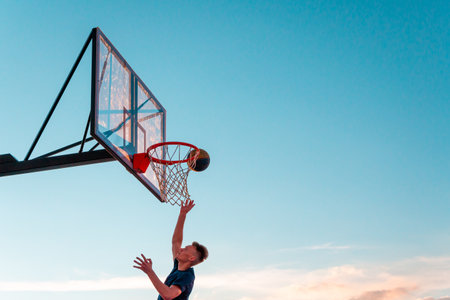 A young basketball player throws a ball into a basketball basket against the blue sky. Transparent plastic basketball shield on the outdoor playground.のeditorial素材