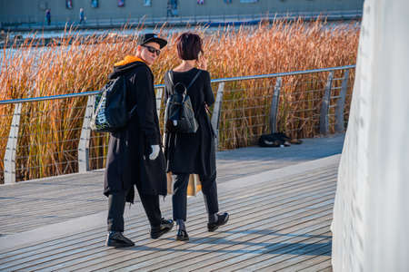 Kazan, Russia - October 14, 2020: Beautiful fashionable young guy and girl in black clothes, with backpacks, walking along a modern wooden embankment with a metal fence along the water's edgeのeditorial素材