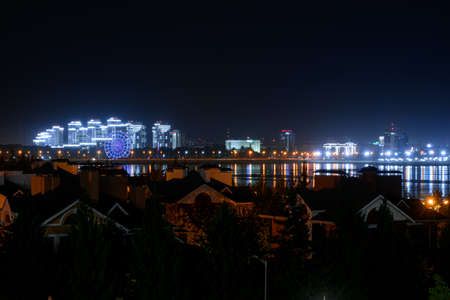 Illuminated buildings and lanterns on the river bank, the multicolored lights of the night city reflected in the water. Ferris wheel on the embankment.の写真素材