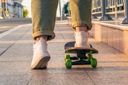Close-up of a woman's legs in jeans and white sports shoes on a skateboard on a city street at...