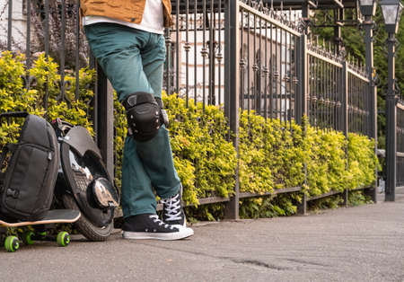 An unrecognizable man stands at a metal fence on a city street. Nearby there is an electric monowheel, a backpack and a skateboard.の写真素材