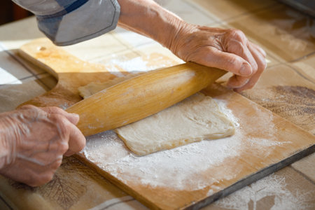 An elderly woman rolls out the dough in flour on a cutting board with a rolling pin. Woman's hands...
