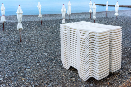Stacked deck chairs on a pebble beach. Closed beach umbrellas. Closing of the beach season. The tourists have left.の写真素材